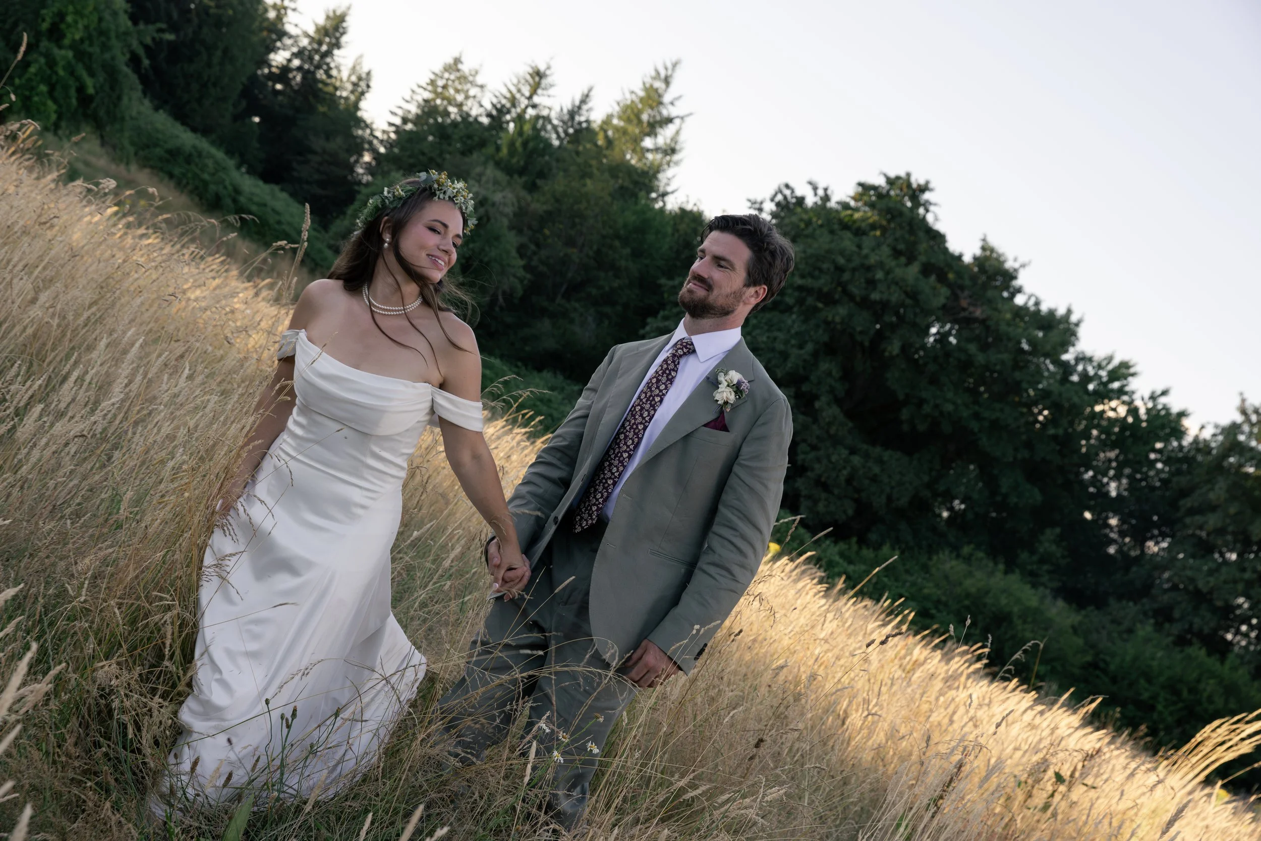 A bride and groom holding hands and walking through a field of tall grass, with trees in the background during sunset. Golden Hour wedding photos. Oregon wedding photographer