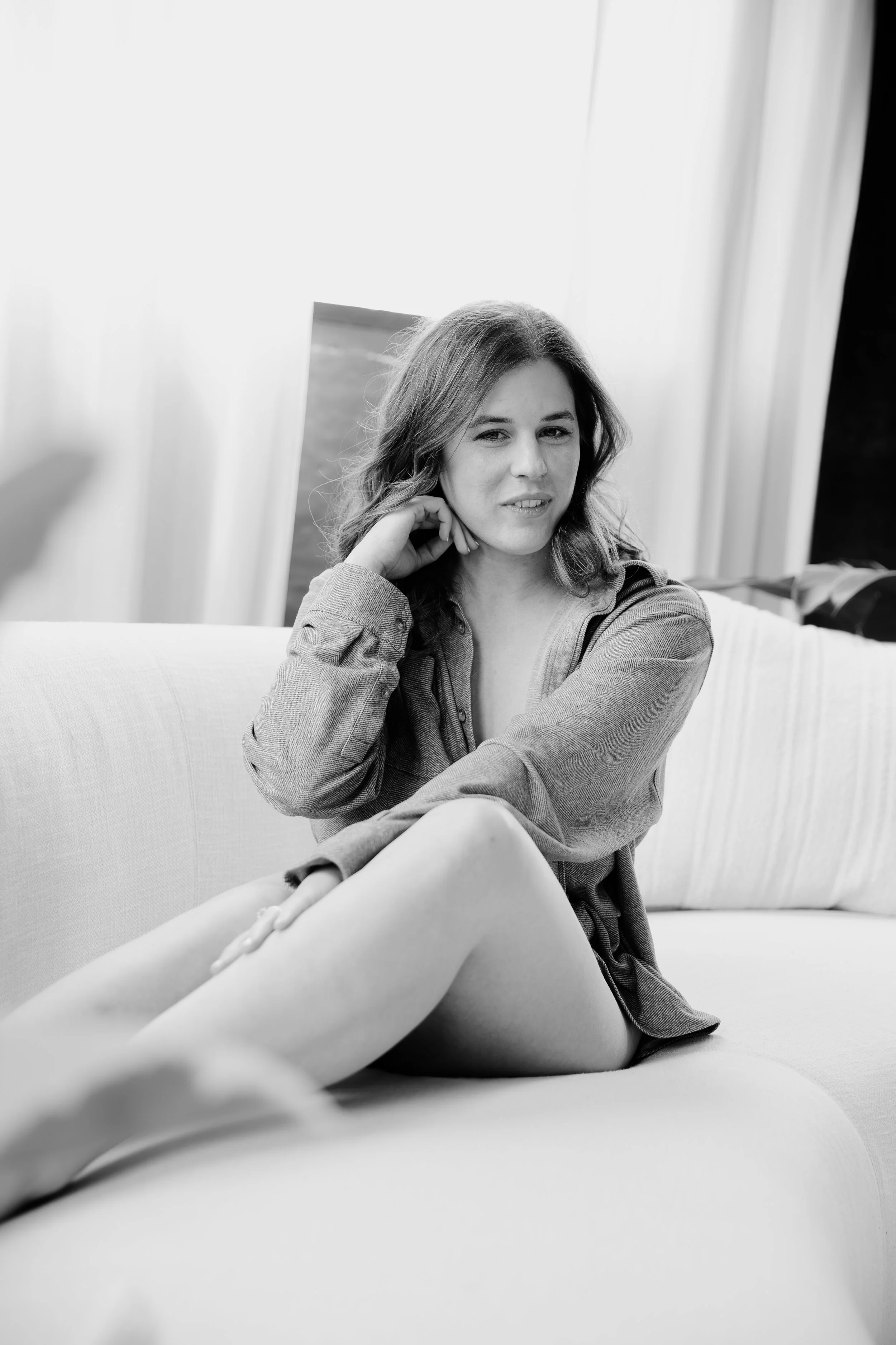 Black and white photo of a woman sitting on a sofa in front of an open window with curtains, looking at the camera with a relaxed expression, and her hair in loose waves.