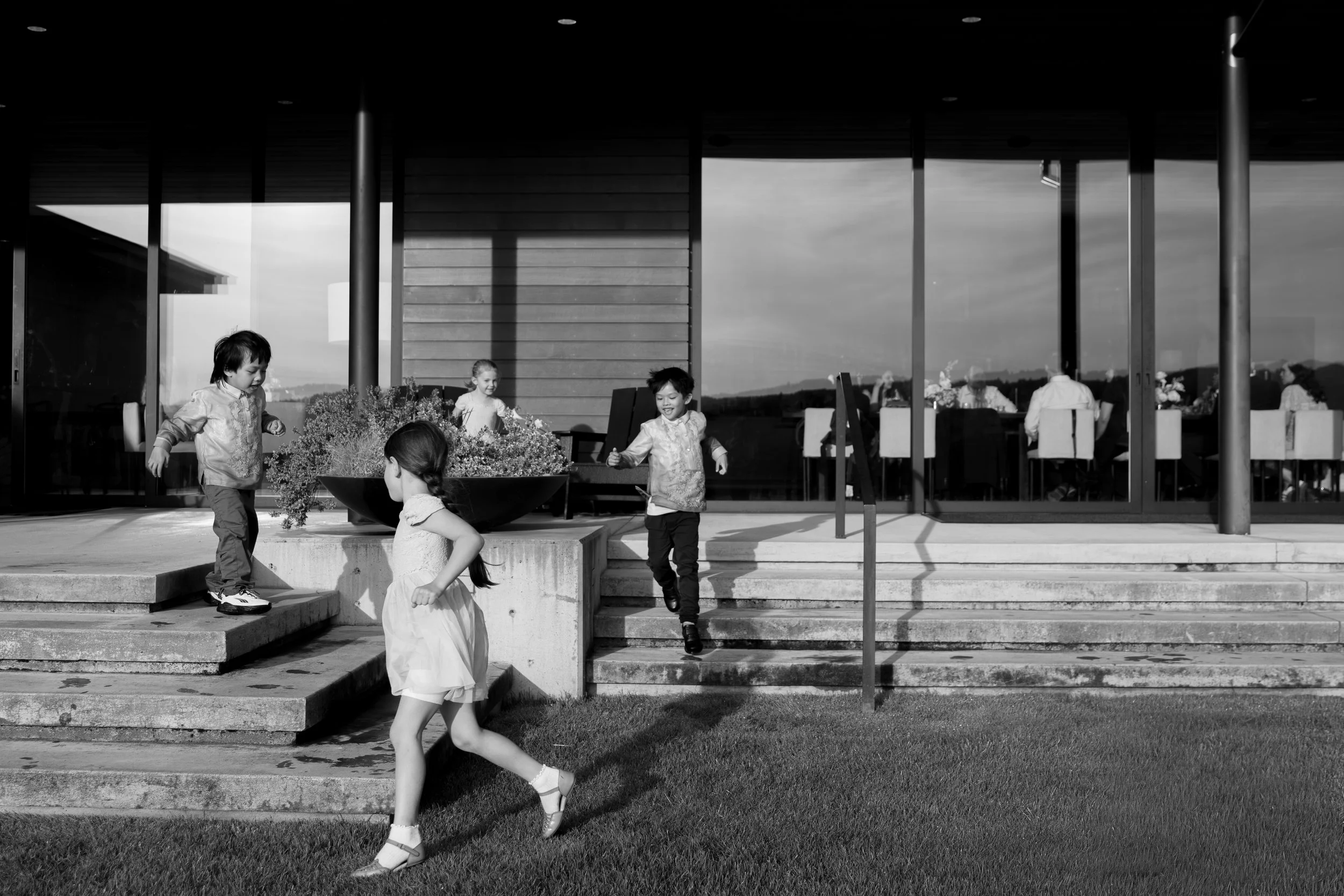 Children playing and running on outdoor stairs near a modern building with large windows, with some people sitting inside visible through the glass.
