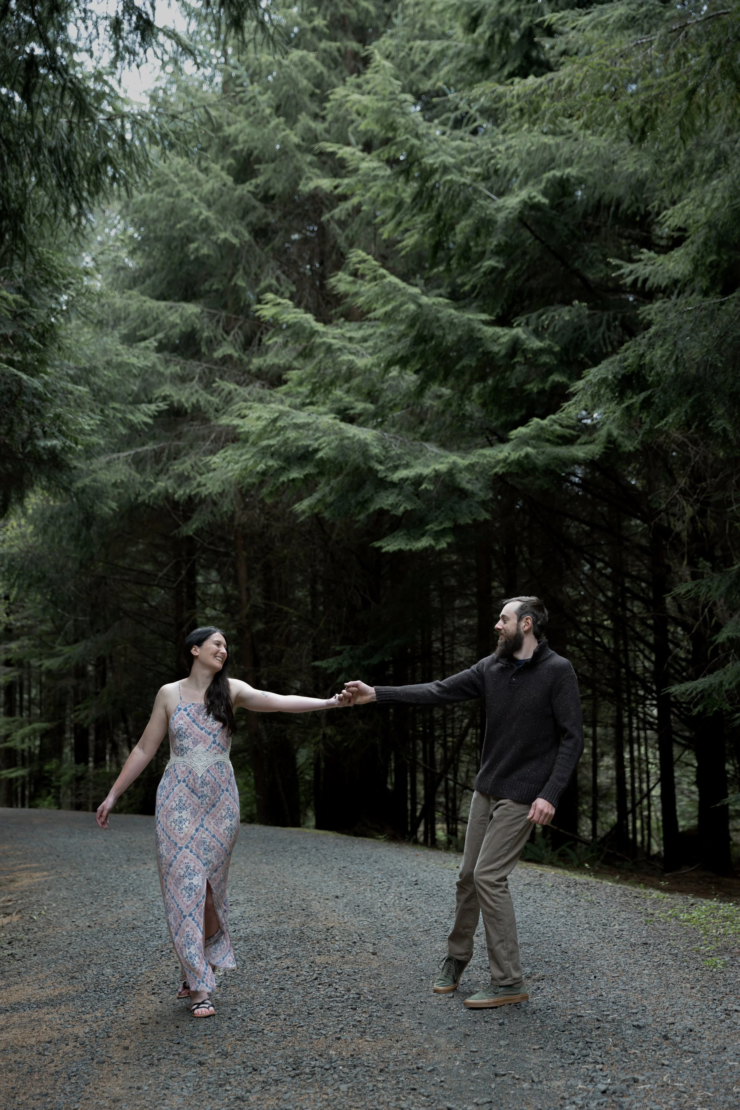 A man and woman holding hands and smiling at each other in a forested area. The woman is wearing a long, patterned dress and the man is dressed in a dark sweater and khakis.