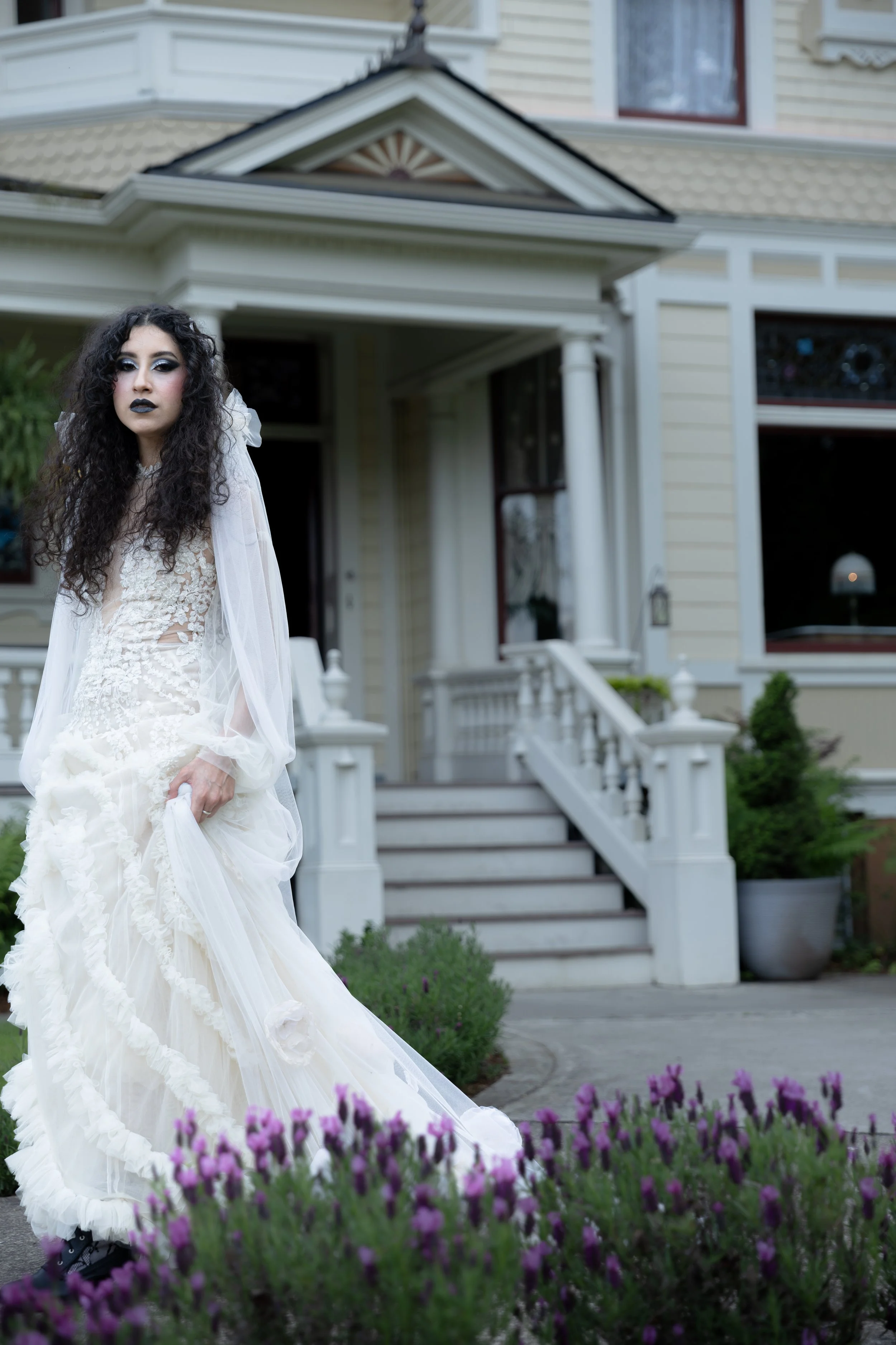A woman with long curly hair wearing dark lipstick and dramatic eye makeup in a white wedding dress standing in front of a house with steps and a porch,. Luxury Oregon Wedding Photographer. Wedding Inspo. Elevated Moody Editorial