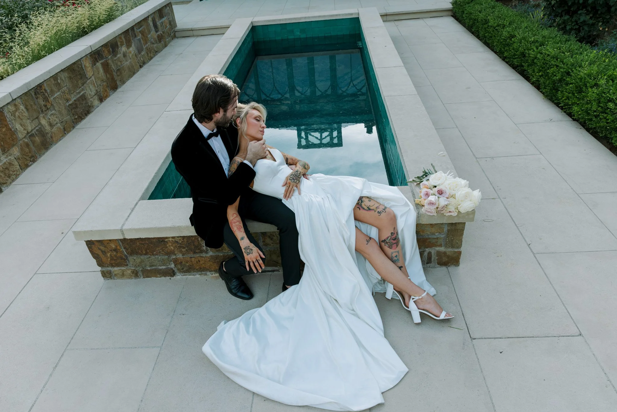 A man and a woman in wedding attire sitting beside a small pool, with the woman resting her head on the man's shoulder.Chateau wedding Bay Area Wedding California Wedding Oregon Wedding Pacific Northwest Wedding White dress Bridal Portraits editorial