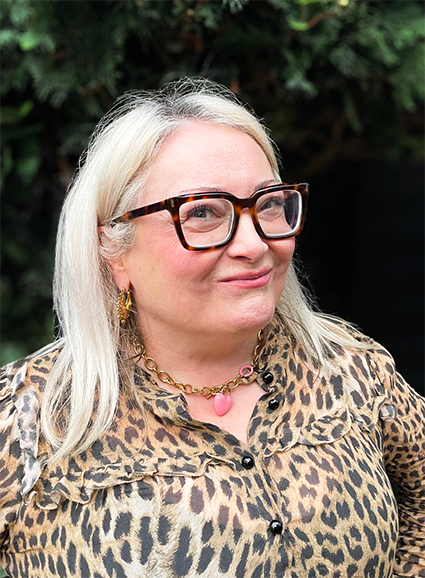 A woman with long blonde hair, wearing glasses, gold jewelry, and a leopard print blouse, smiling outdoors with green foliage in the background.