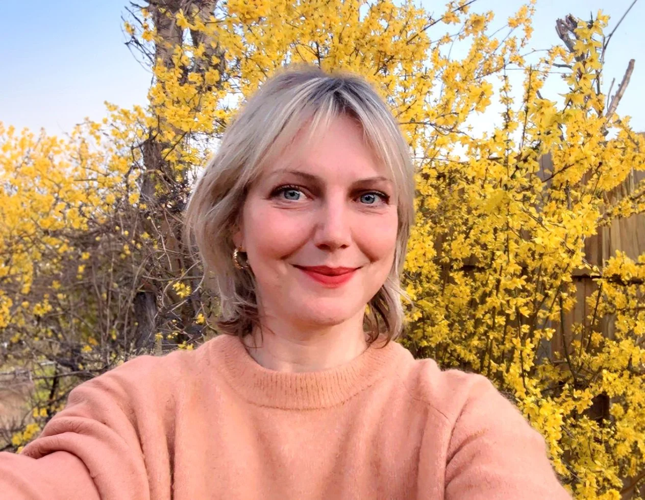 A woman taking a selfie outdoors in front of a yellow flowering tree, wearing a peach-colored sweater and earrings, with a fence in the background.