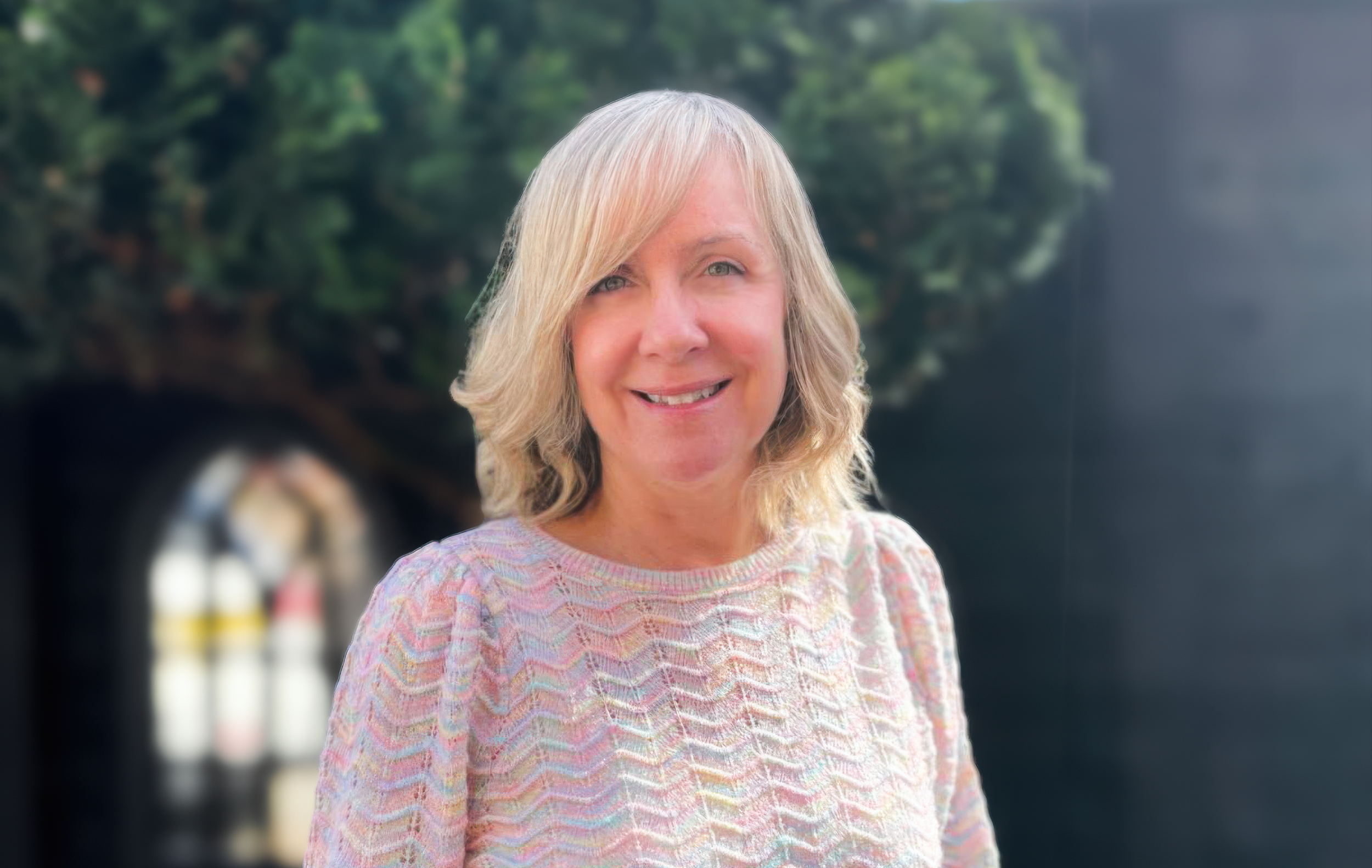 Middle-aged woman with blonde hair smiling, wearing a colorful knit sweater, outdoors with green foliage in the background.