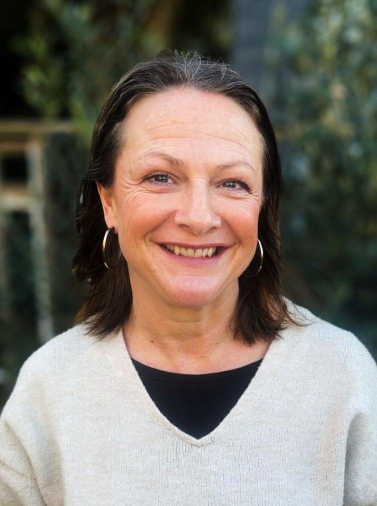 A woman with shoulder-length dark hair, wearing hoop earrings and a light-colored sweater, smiling outdoors with a blurred background of trees.