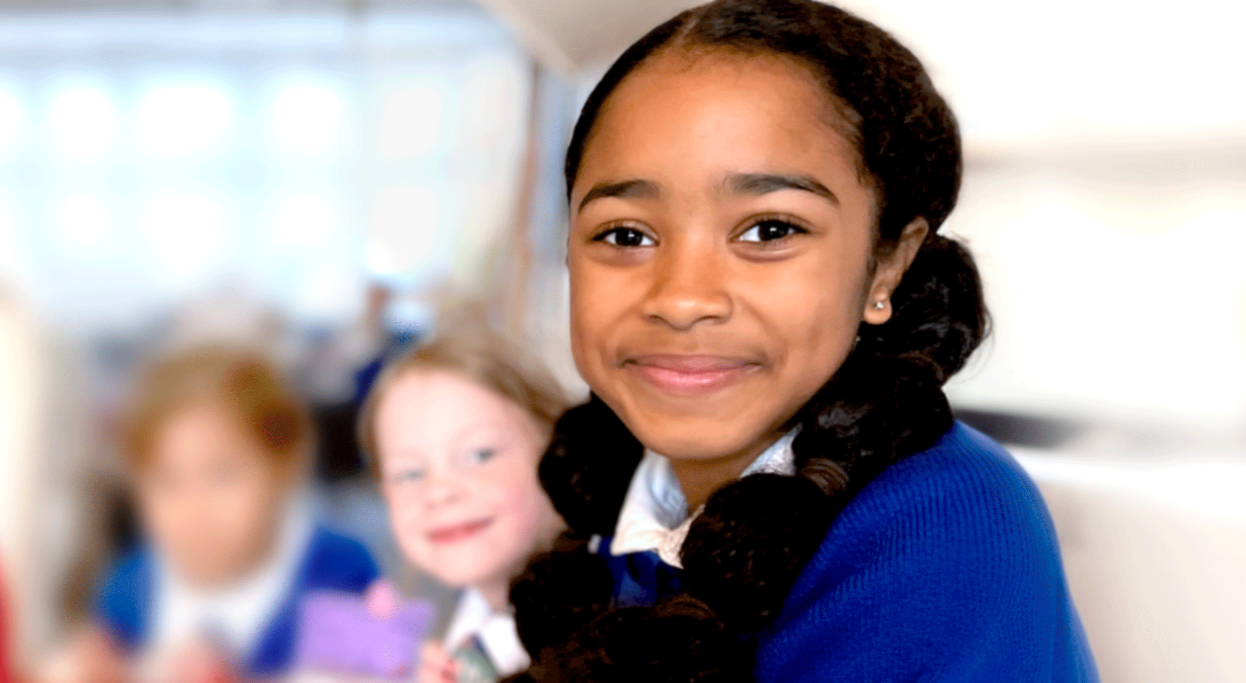 Close-up of a smiling African American girl with dark curly hair and a blue sweater, in a classroom with other children in the background.