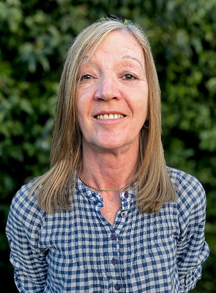 A middle-aged woman with blonde hair and a blue checkered shirt standing outdoors in front of greenery.