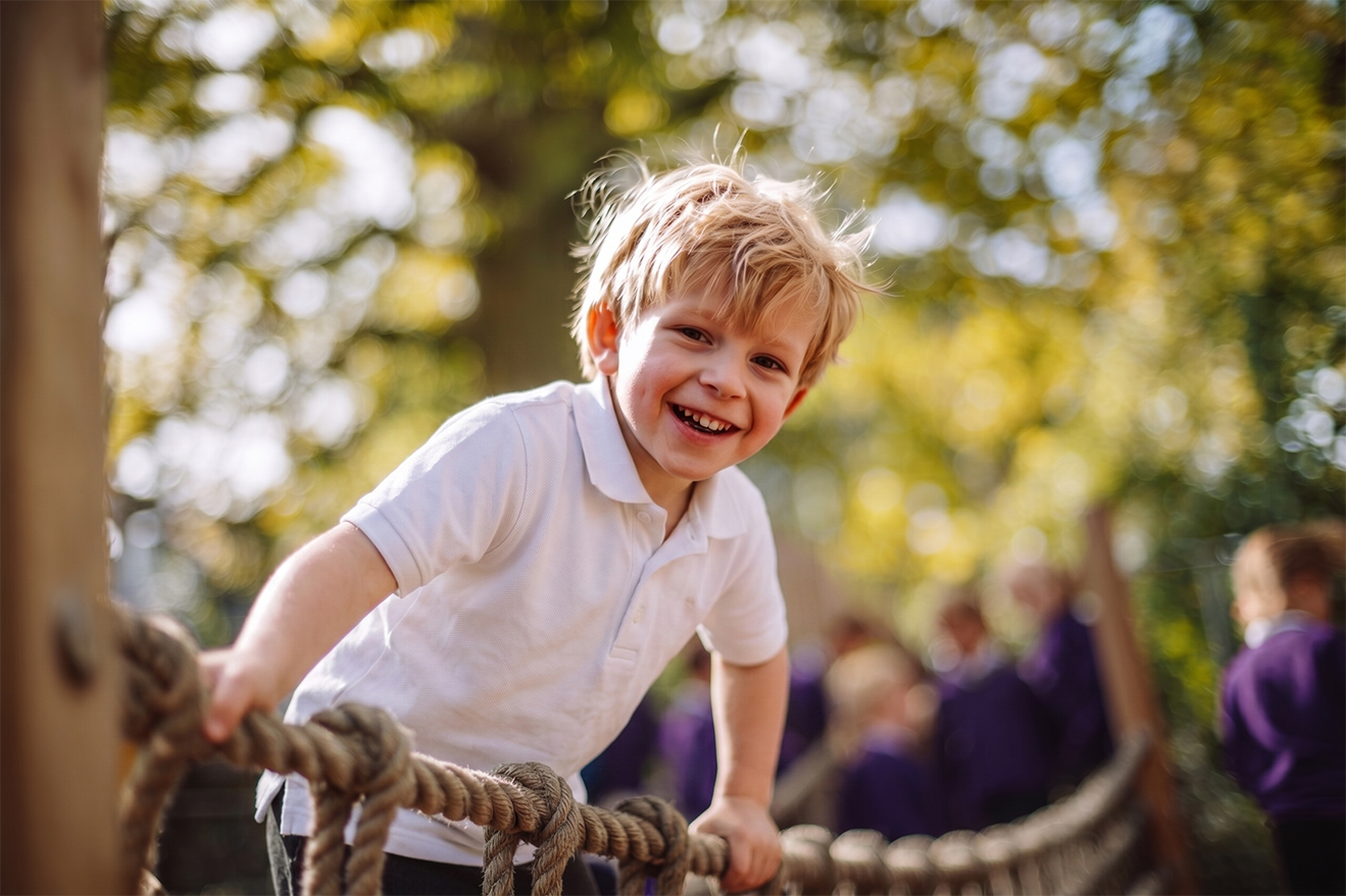 A young boy with blonde hair and a white shirt smiling while climbing on a ropes bridge outdoors, with blurred children in purple uniforms in the background and trees with sunlight filtering through.