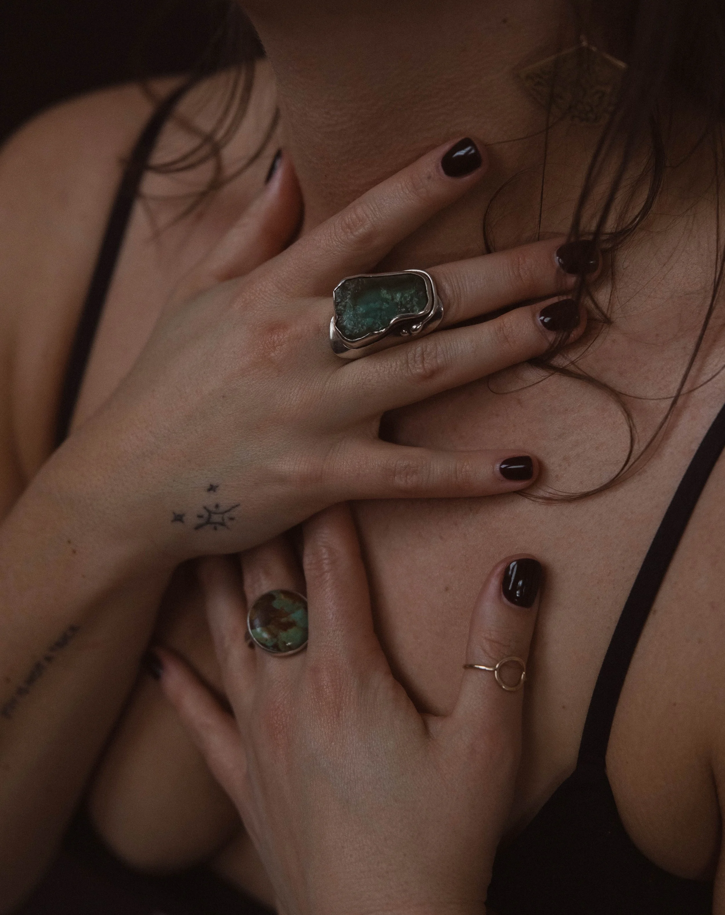 Close-up of two hands with rings, touching skin and hair. One hand has a large silver ring with a green stone, the other hand has a silver ring with a round green stone. Both hands have dark nail polish. One hand has a small tattoo of a symbol with stars.