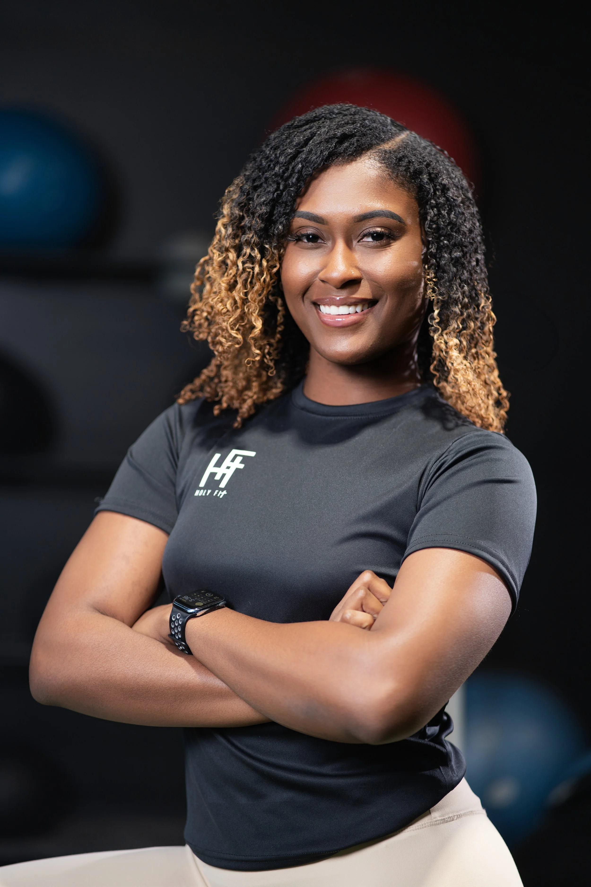Smiling woman with curly hair in a black fitness shirt crossing her arms, wearing a smartwatch.