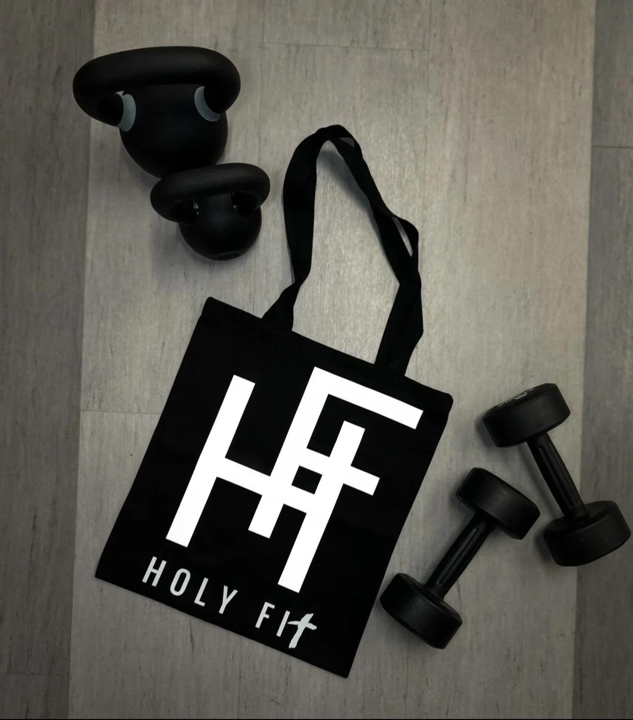 A black tote bag with the words 'Holy Fit' and a stylized cross logo, two black kettlebells, and two small black dumbbells on a floor.