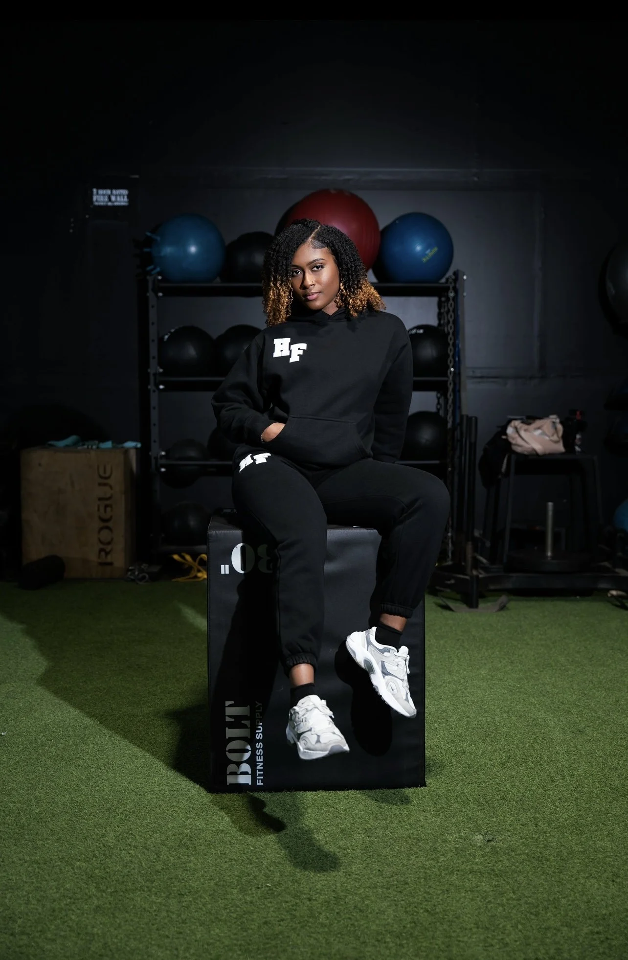Woman in black athletic wear sitting on a gym box with exercise balls in the background.