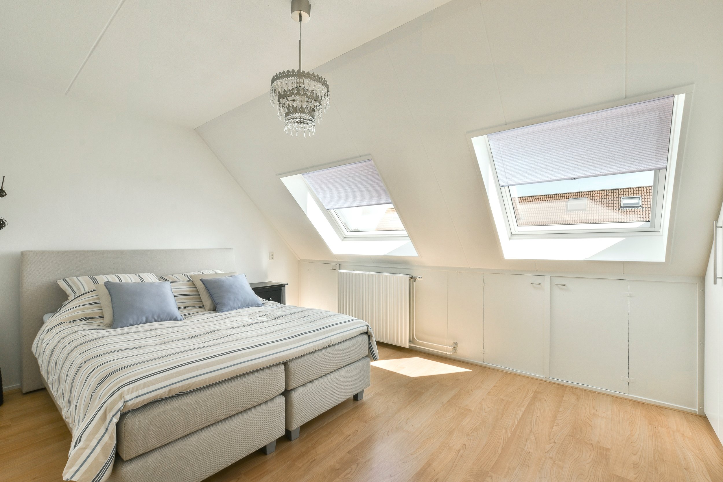 Bright attic bedroom with a double bed, striped bedding, gray pillows, wooden floor, chandelier, and skylight windows.