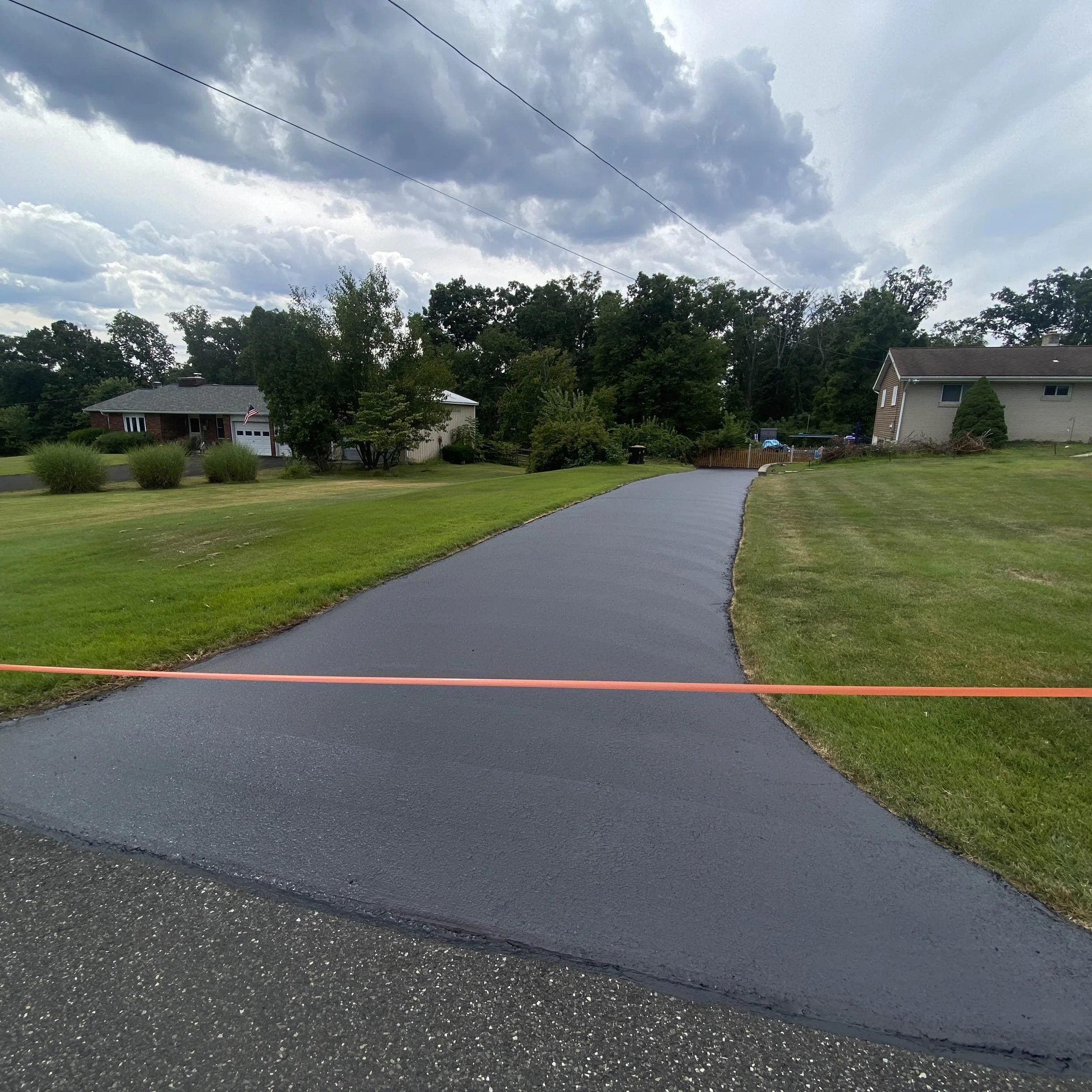 Freshly poured black asphalt driveway in a residential neighborhood with green lawns on both sides, houses in the background, and a cloudy sky overhead.