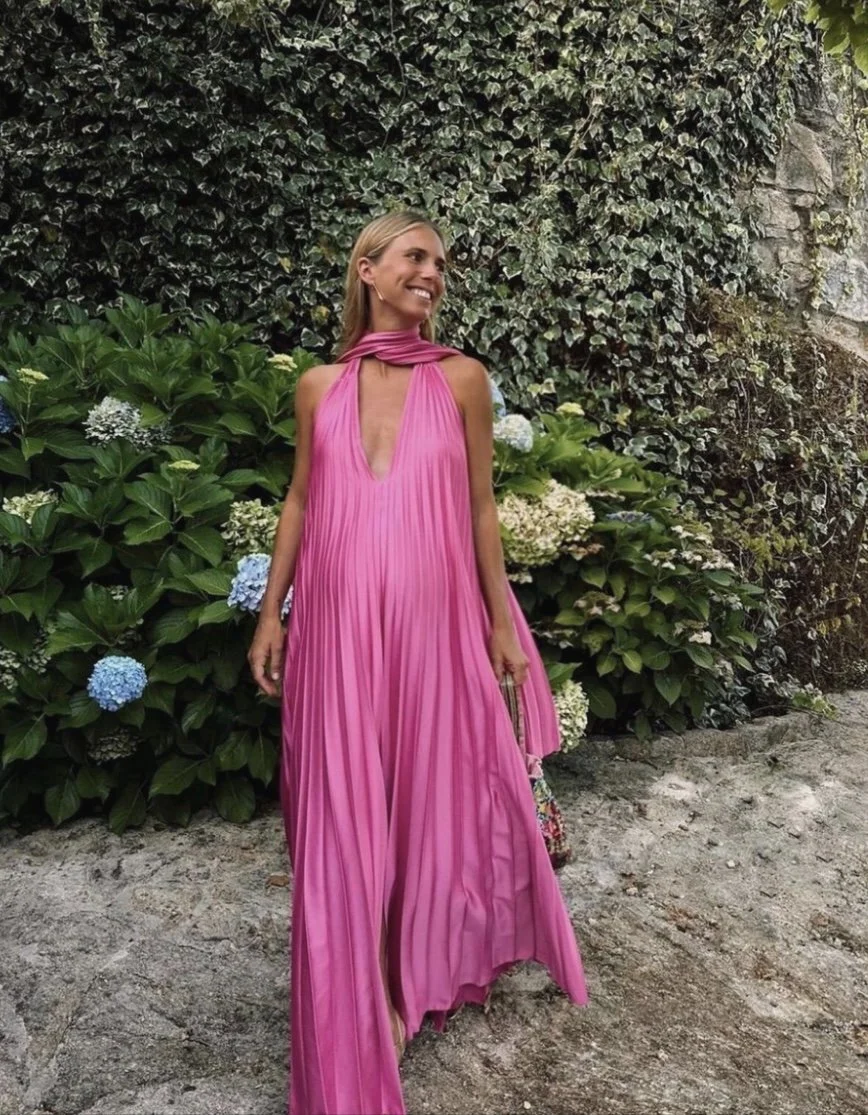 Woman in a bright pink pleated dress standing outdoors in front of green bushes and hydrangea flowers, smiling and looking to her left.