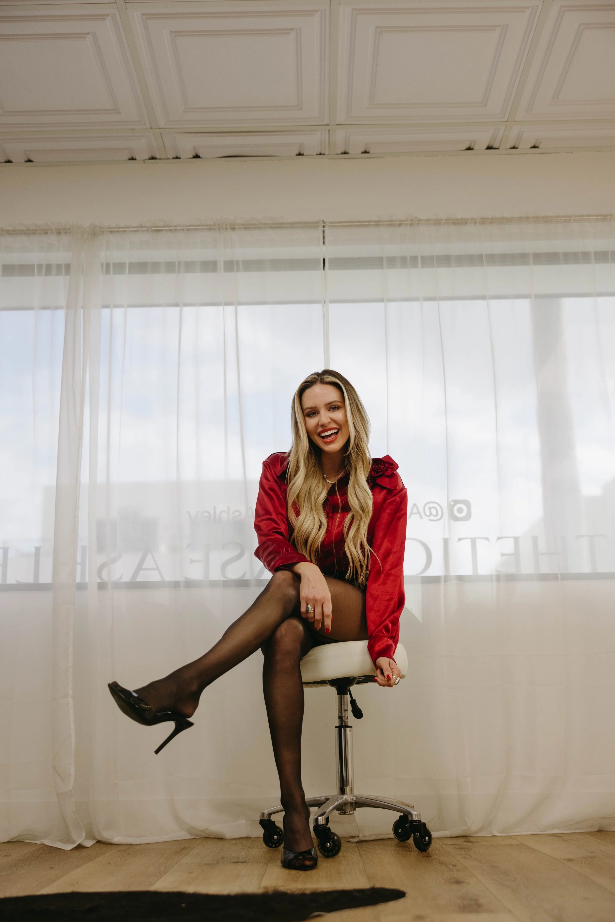 A woman with long wavy hair wearing a red blouse and black stockings, sitting on a white office chair, smiling and posing in front of a window with sheer white curtains.