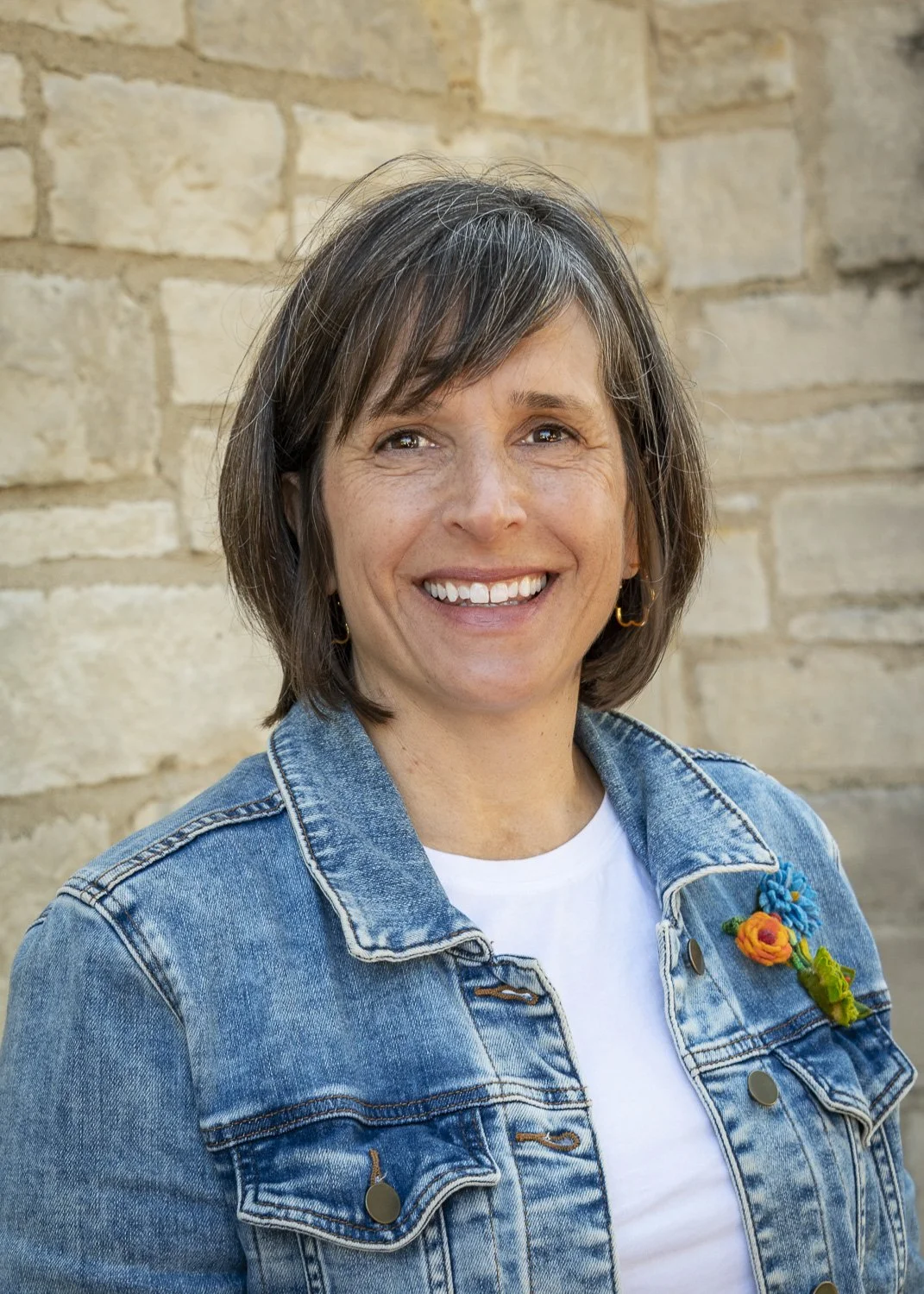Smiling woman with short brown hair, wearing a white short-sleeved blouse and blue jeans, standing outdoors in a garden with green trees and a house in the background.