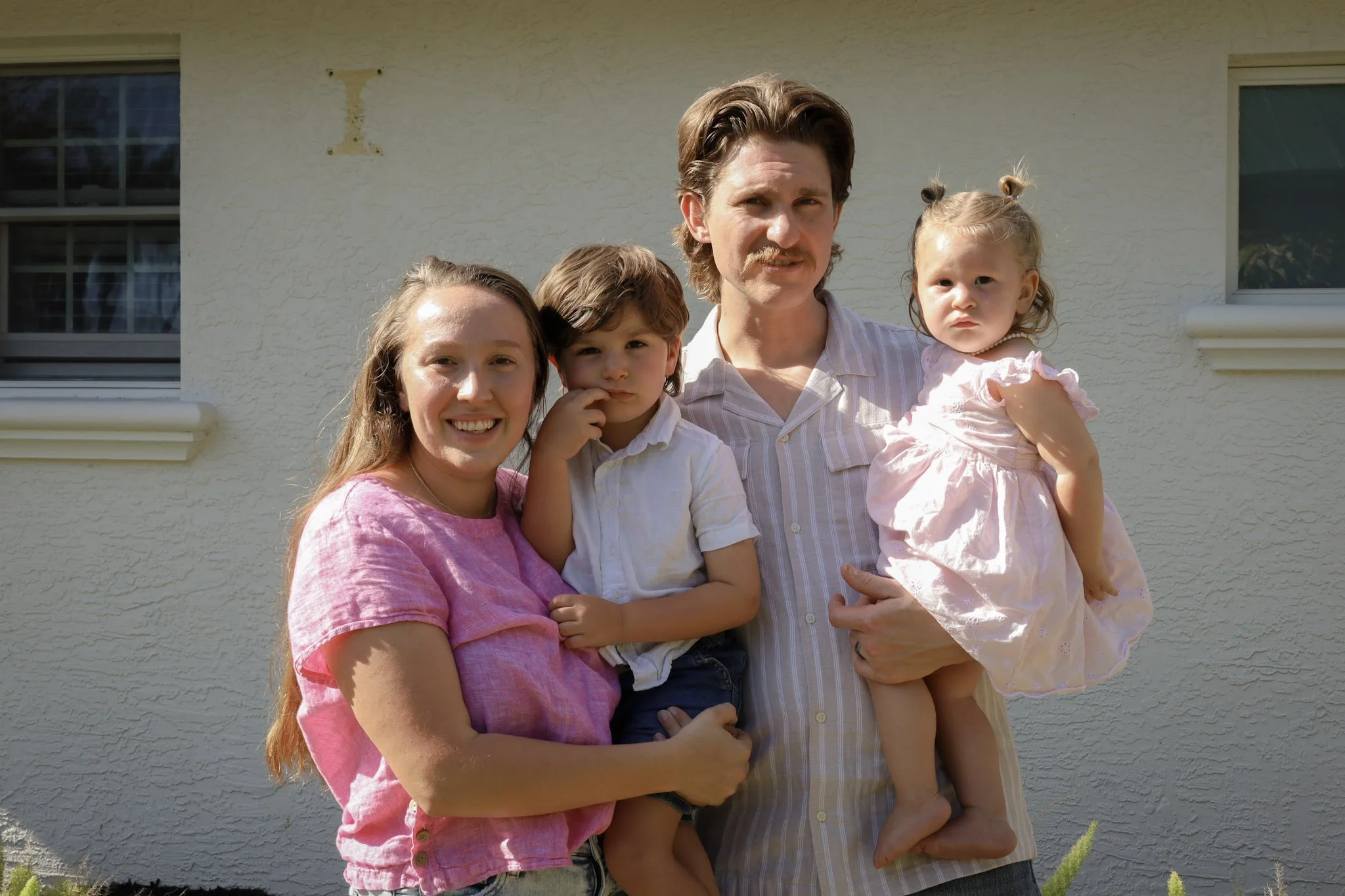 A family of four posing outdoors in front of a beige building with two windows, smiles, and casual clothing. The parents are holding their children, a boy and a girl.
