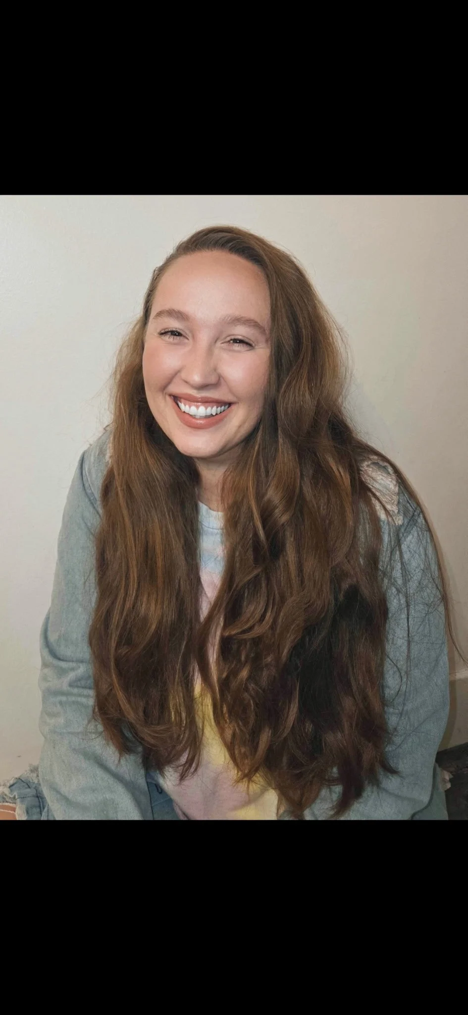 Young woman with long wavy brown hair smiling at the camera, wearing a denim jacket and a pastel-colored shirt, against a plain light background.