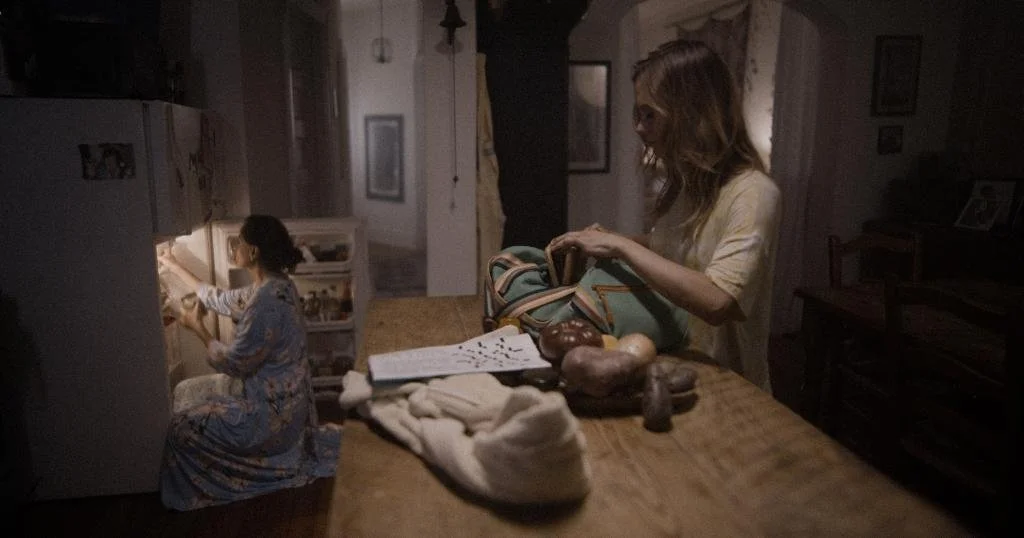 A woman with blonde hair packing a bag at a kitchen table while a girl in pajamas looks inside a refrigerator in a dimly lit room.