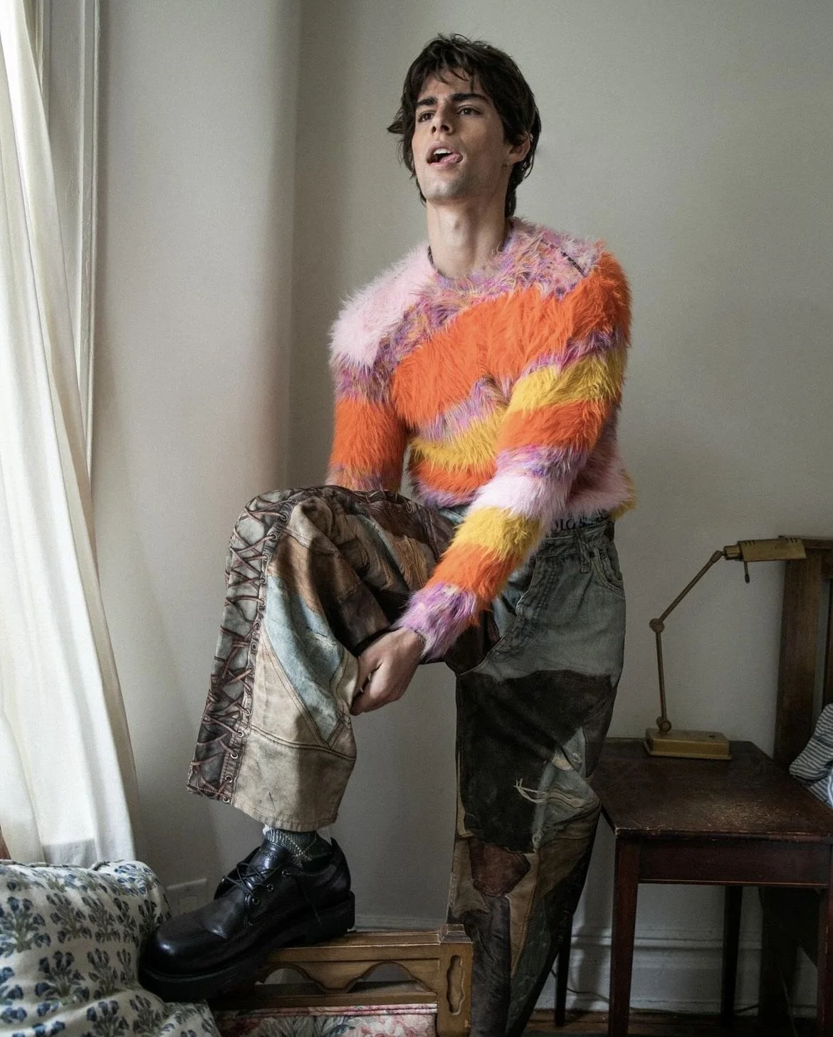 A person wearing a colorful, fluffy sweater standing on a chair, holding a pair of patchwork shorts, with a room and a small desk lamp in the background.