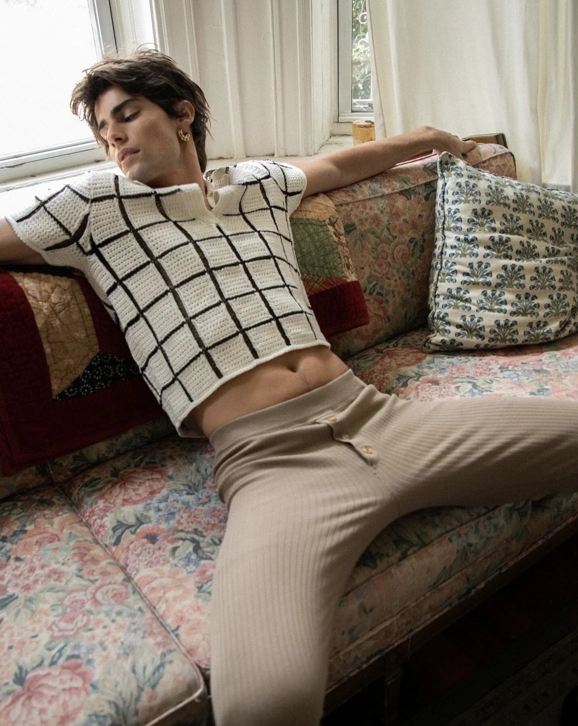 A young person with short dark hair and gold earrings is lounging on a floral-patterned sofa, wearing a white cropped top with black grid lines and beige high-waisted pants, with arms outstretched and eyes closed.