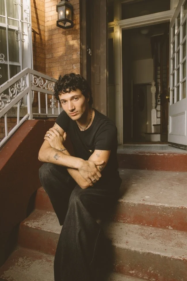 Young man with curly dark hair, tattoos, and jewelry, wearing a black t-shirt and dark pants, sitting on the steps outside a brick building with a white railing and an open door, illuminated by a wall-mounted lantern.
