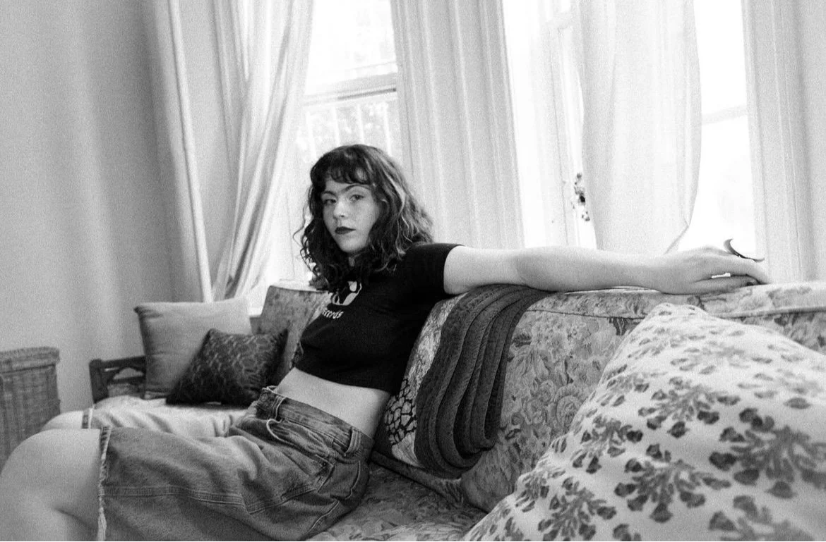 A young woman with curly dark hair and dark lipstick sitting on a floral-patterned sofa with her arm extended along the back, wearing a cropped black t-shirt and high-waisted denim shorts, in a room with large windows and curtains.