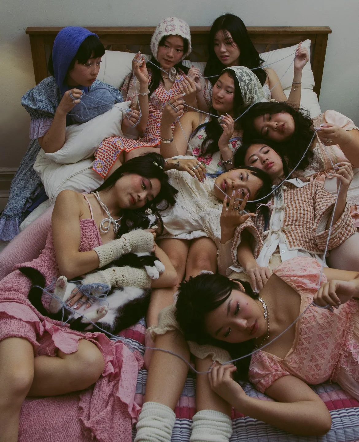 A group of women lying on a bed, holding yarn and knitting; some women are wearing pajamas, and there is a black and white cat among them.