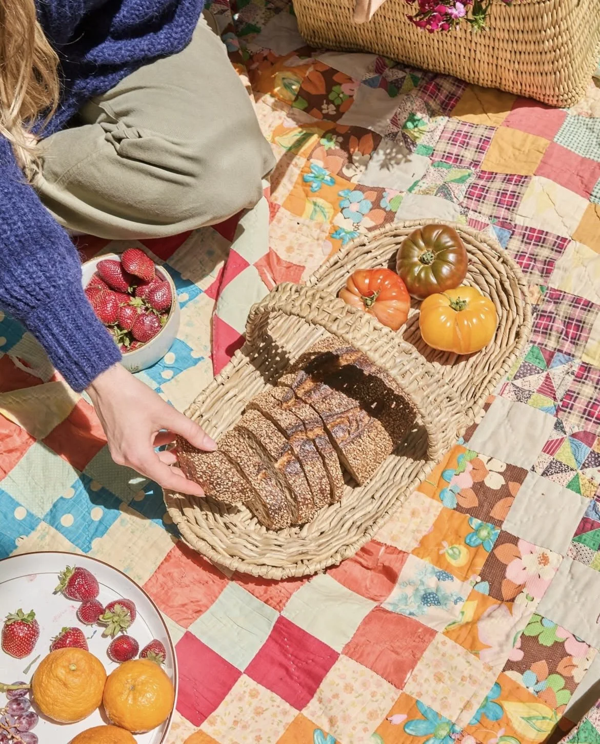 A picnic scene with a quilted blanket, a person cutting bread, a basket with heirloom tomatoes, strawberries, and oranges, and other mixed fruit.