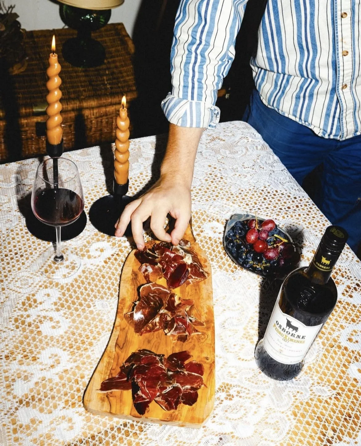 Person in a striped shirt catches slices of cured ham from a wooden serving board on a white lace tablecloth, accompanied by a glass of red wine and a bottle of red wine, with a bowl of grapes and lit candles on the table.