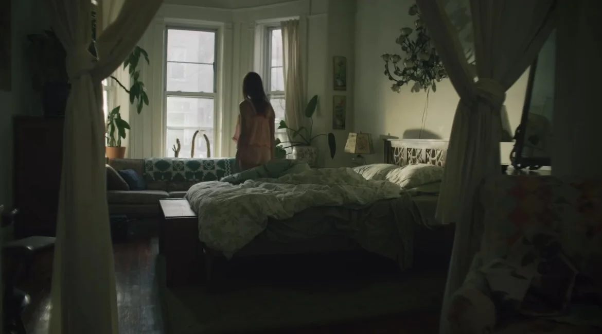 A woman standing by a window in a sunlit bedroom, viewed through curtains, with a bed, plants, and artwork visible in the room.