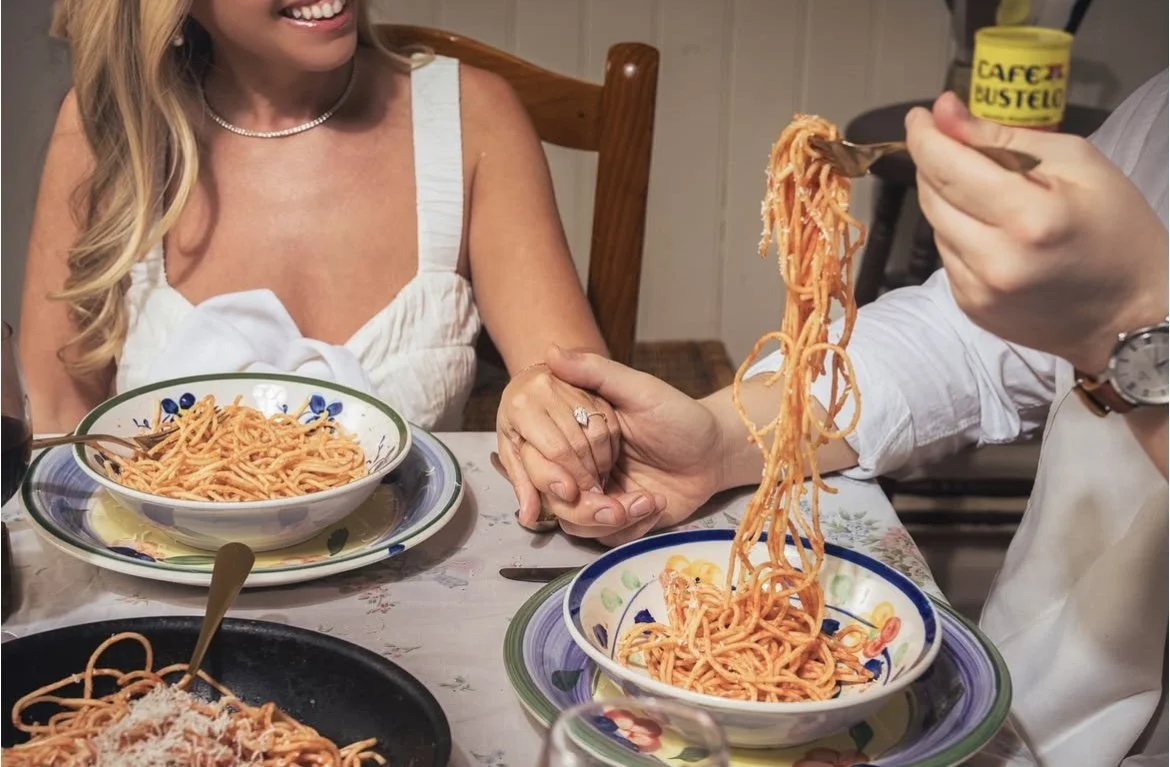 A woman at a dinner table receiving a hand-taken plate of spaghetti from a server, with bowls of spaghetti and a plate of spaghetti topped with grated cheese on the table.
