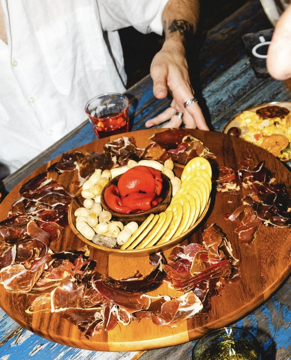 A round wooden platter of assorted cured meats, cheese slices, and a small bowl of red peppers, with a glass of red wine next to it on a rustic wooden table.