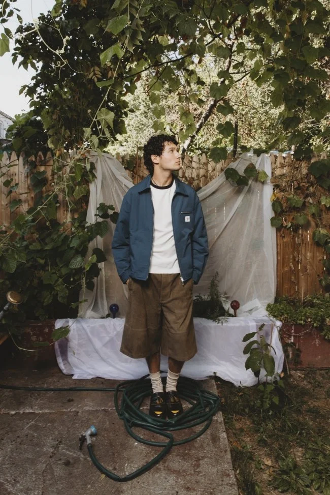 A young man with curly hair and light skin stands outdoors in a garden, surrounded by green plants and vines. He is wearing a blue jacket, a white shirt, brown wide-leg shorts, and striped socks with sandals. Behind him, there is a makeshift backdrop
