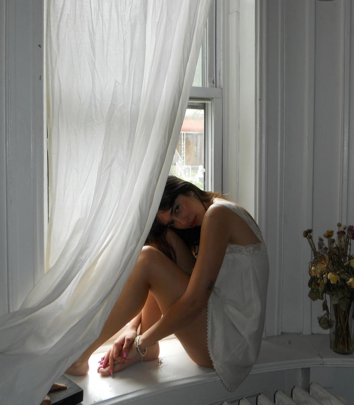 A woman sitting on a windowsill inside a room with white wooden walls, partially covered by a white sheer curtain, with a vase of dried flowers nearby.