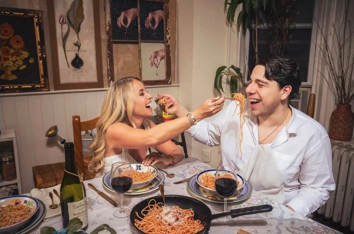 A couple sharing a fun moment at dinner, with spaghetti. The woman is feeding the man with spaghetti, both smiling and laughing, seated at a table with wine glasses, a bottle of wine, and plates of spaghetti.
