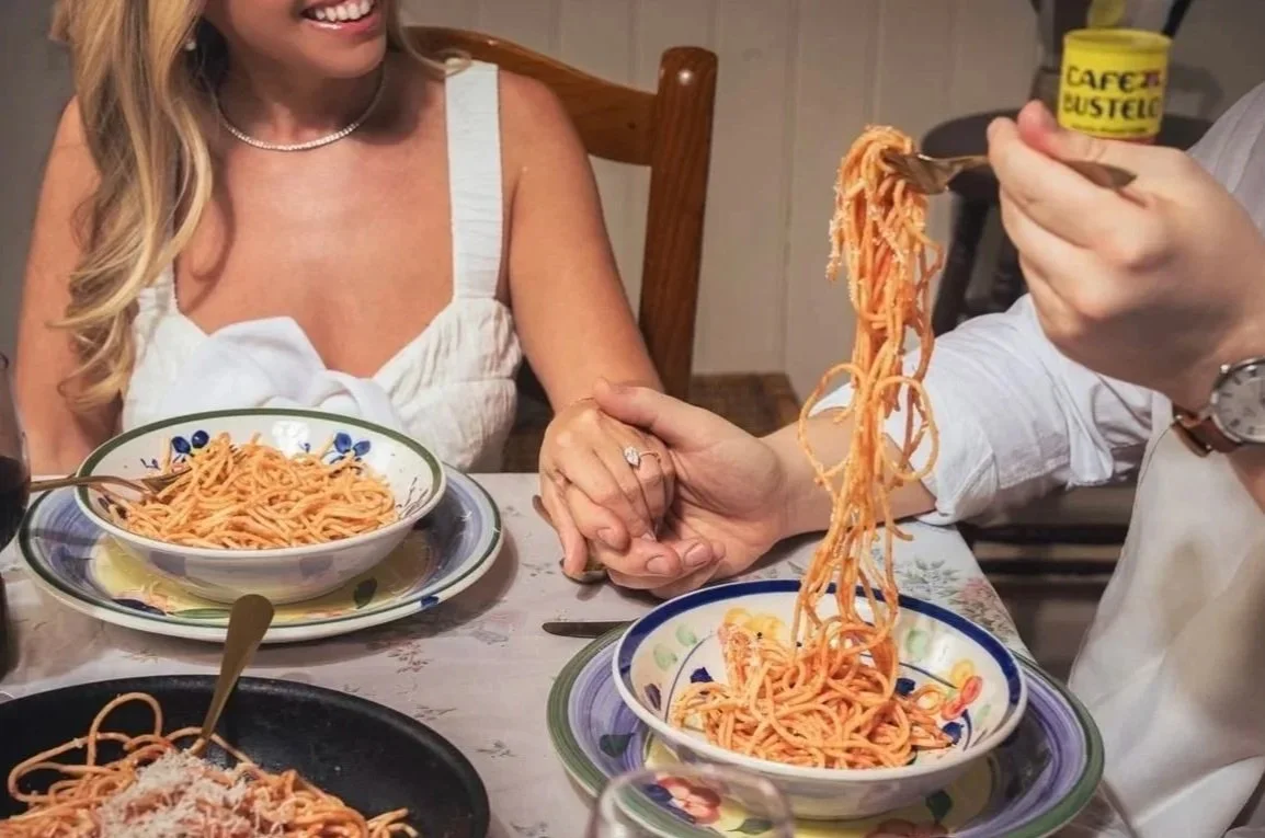 A woman at a dinner table receiving a hand-taken plate of spaghetti from a server, with bowls of spaghetti and a plate of spaghetti topped with grated cheese on the table.