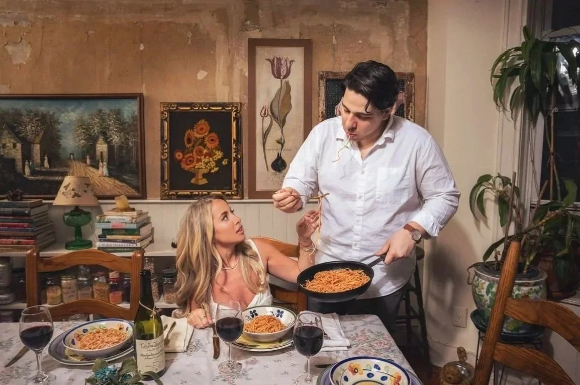 A man serving spaghetti to a woman at a dining table with glasses of red wine, set in a cozy room with artwork and plants.