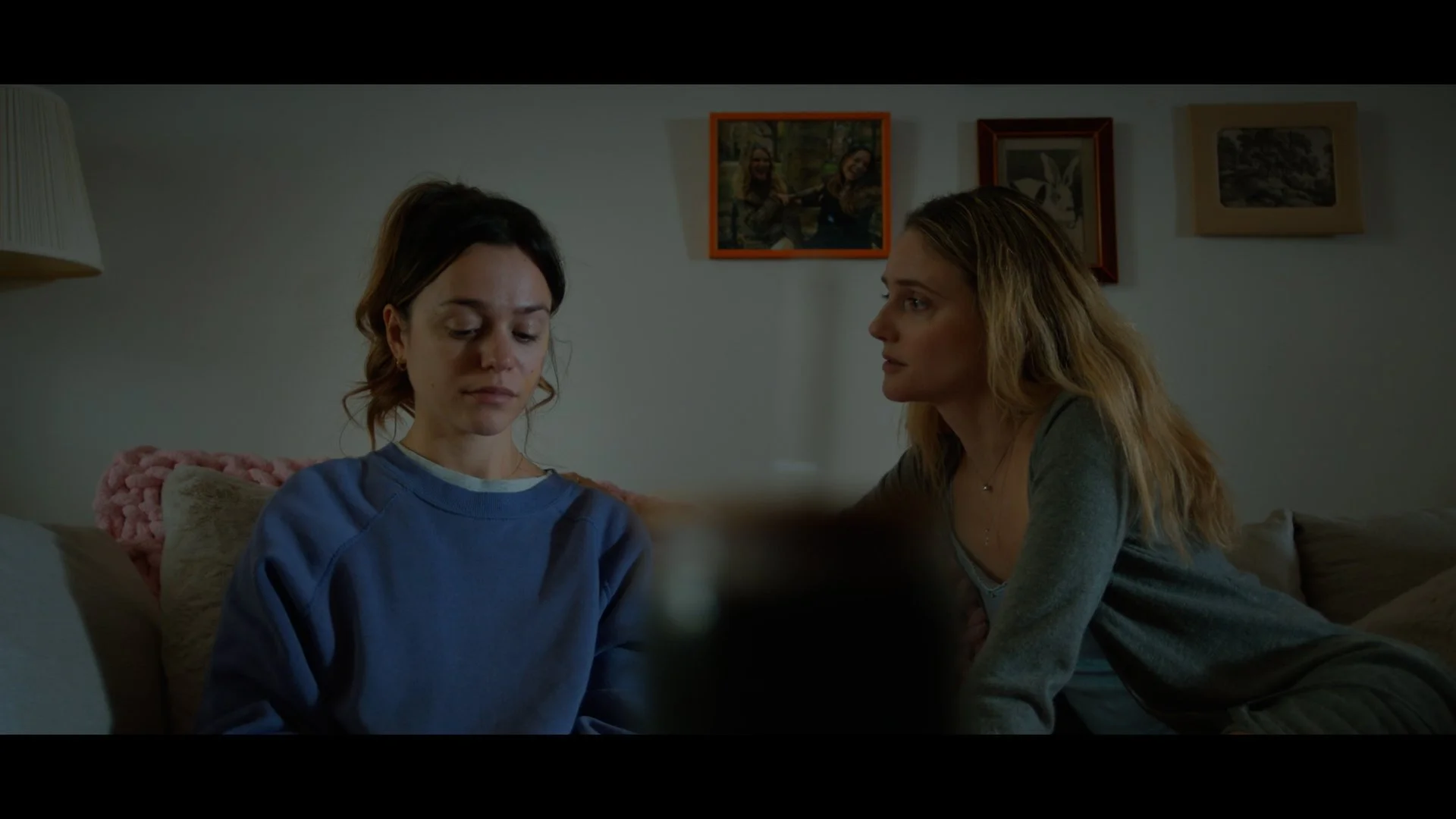 Two women sit on a couch in a dimly lit room, one with dark hair looking sad and a woman with blonde hair speaking to her. Framed pictures hang on the wall behind them.