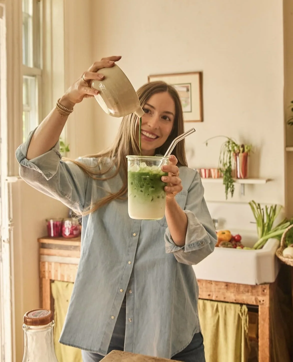 A woman in a light blue shirt making a matcha latte, pouring soy milk into a clear glass with a green drink inside, in a cozy kitchen setting.