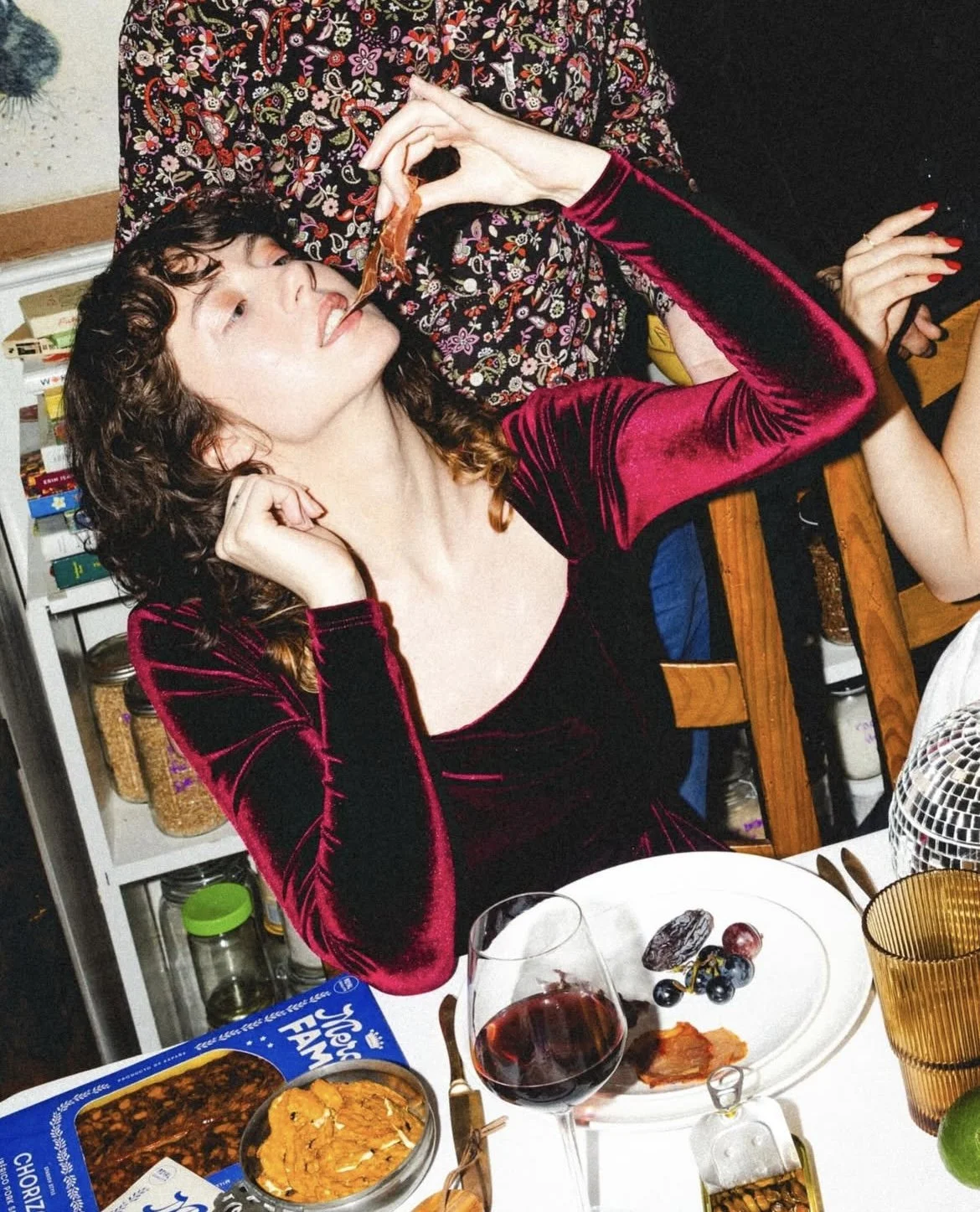 A woman in a red velvet dress is sitting at a table with food and drink, and someone is feeding her a piece of food using chopsticks.