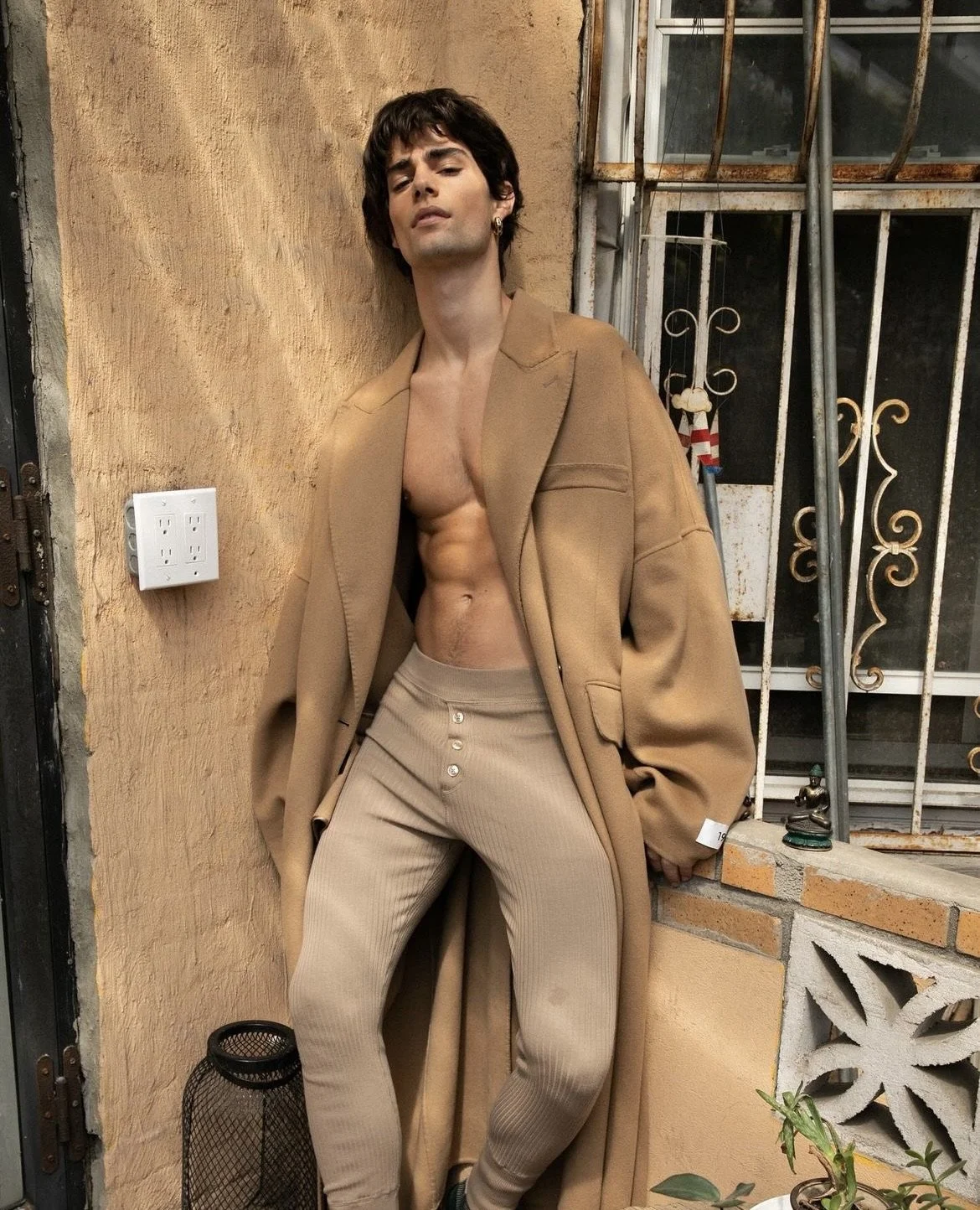 A young man with dark hair and earrings leans against an outdoor wall, wearing a tan oversized coat and beige ribbed pants with buttons, exposing his bare chest and abs.