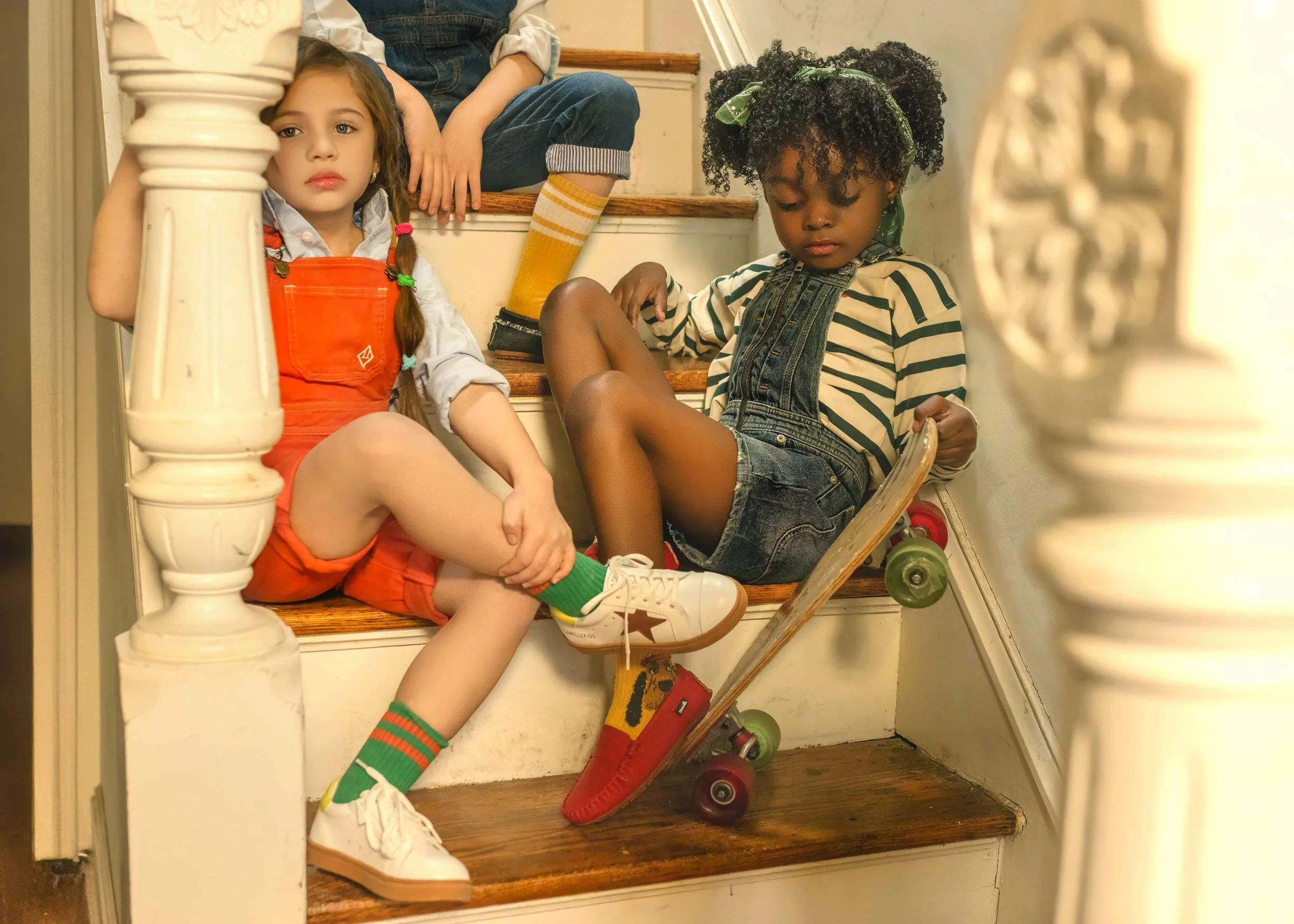 Three children sitting on a carpeted staircase. A girl with long brown hair and colorful hairbands is sitting with her leg crossed, wearing orange overalls, a white shirt, and white sneakers with green and red socks. A boy with curly hair tied with a