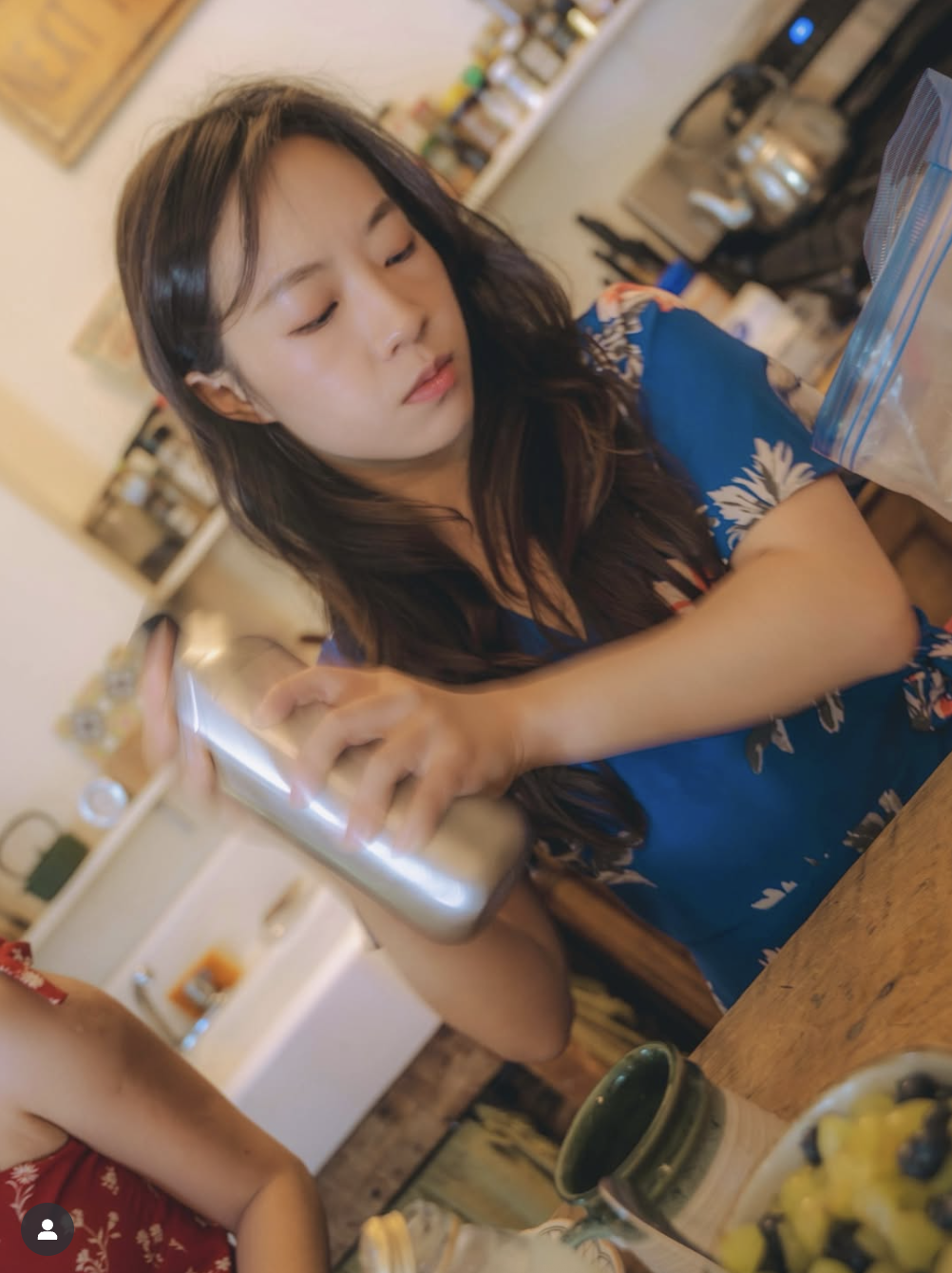A young woman with long dark hair, wearing a colorful short-sleeved dress, is looking down while adjusting her hair or head with her left arm resting on a wooden table. The background shows shelves with jars and kitchen items, indicating a home kitch