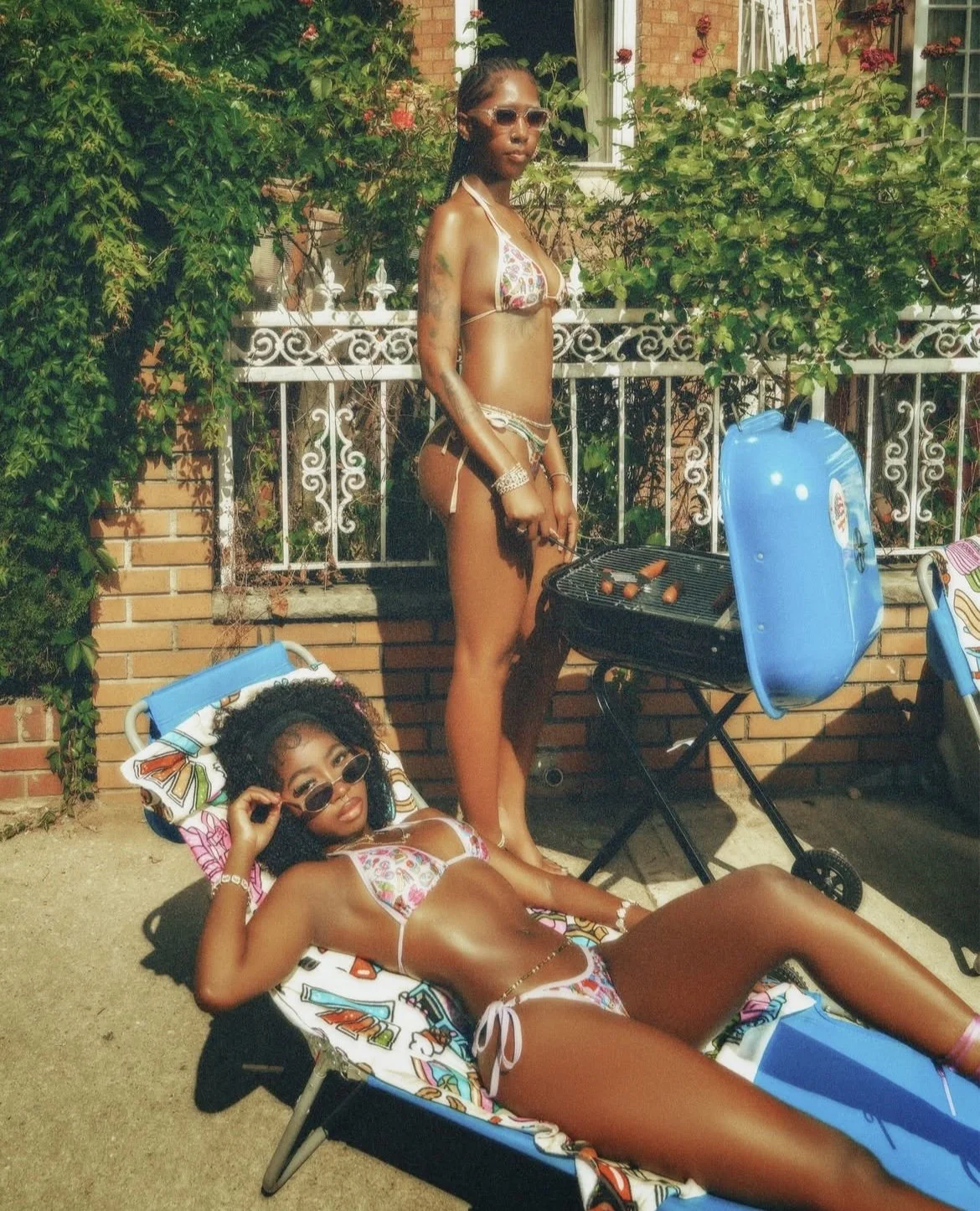 Two women in bikinis relaxing outdoors, one lying on a lounge chair and the other standing beside a barbecue grill, with a garden and brick wall in the background.