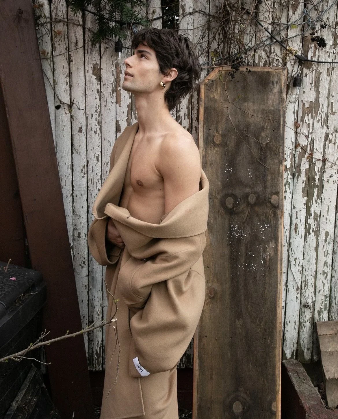 A young man with dark, tousled hair, wearing earrings, is standing shirtless outdoors against a weathered wooden fence. He is partially wrapped in a beige jacket and matching pants, holding the jacket closed with one hand, and looking upward and to t