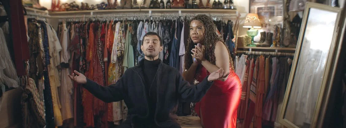 A man and woman in a vintage clothing store with racks of dresses and garments behind them. The man has short dark hair and is wearing a black jacket, sitting with his arms outstretched, eyes closed, and mouth slightly open. The woman has long curly 