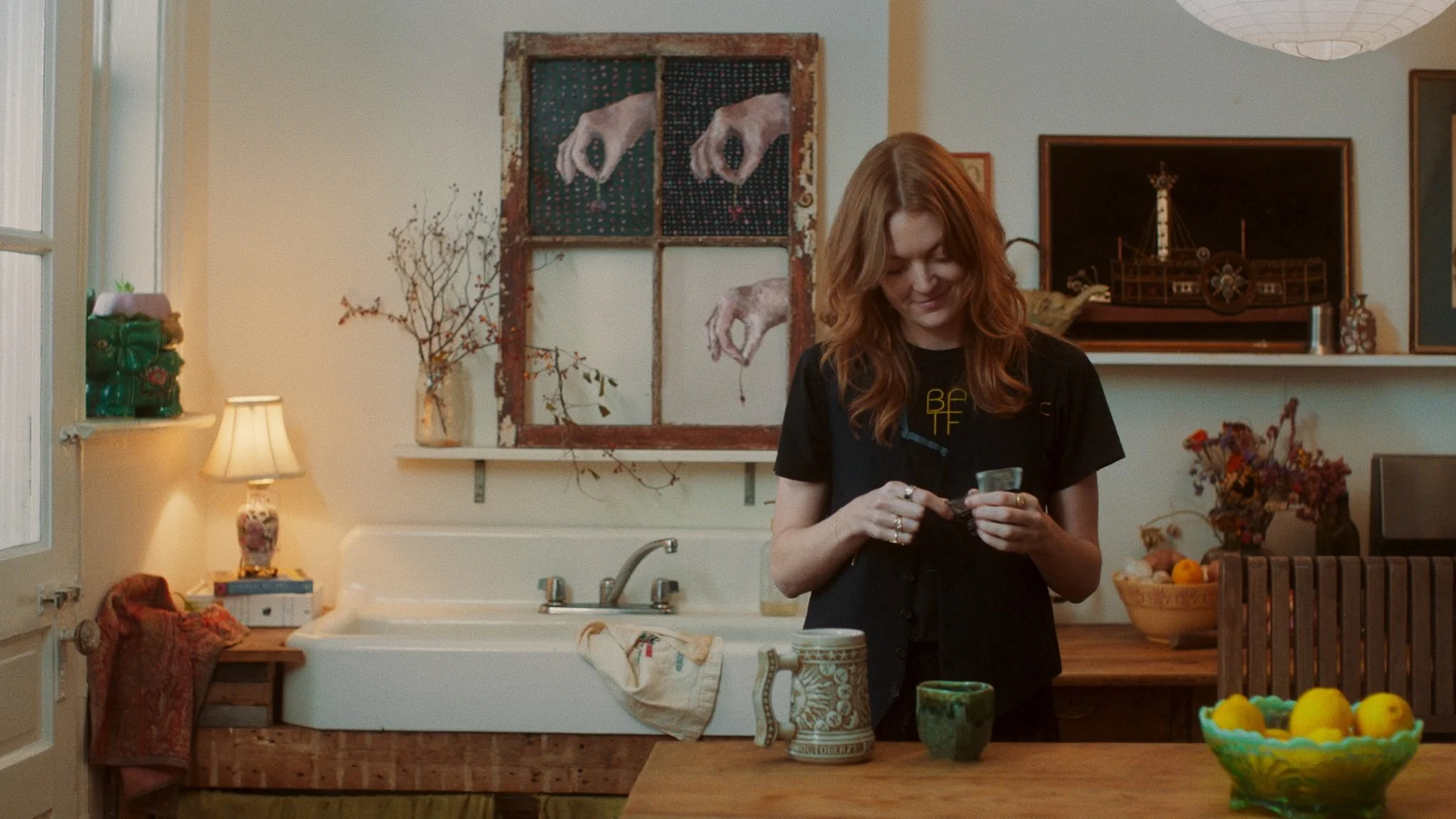 A woman with red hair smiling in a cozy kitchen. She is holding a small object, possibly a jar or a cup. The kitchen has a white sink with a vintage faucet, a wooden table with a ceramic pitcher, a green cup, and a bowl of lemons. There are decorativ